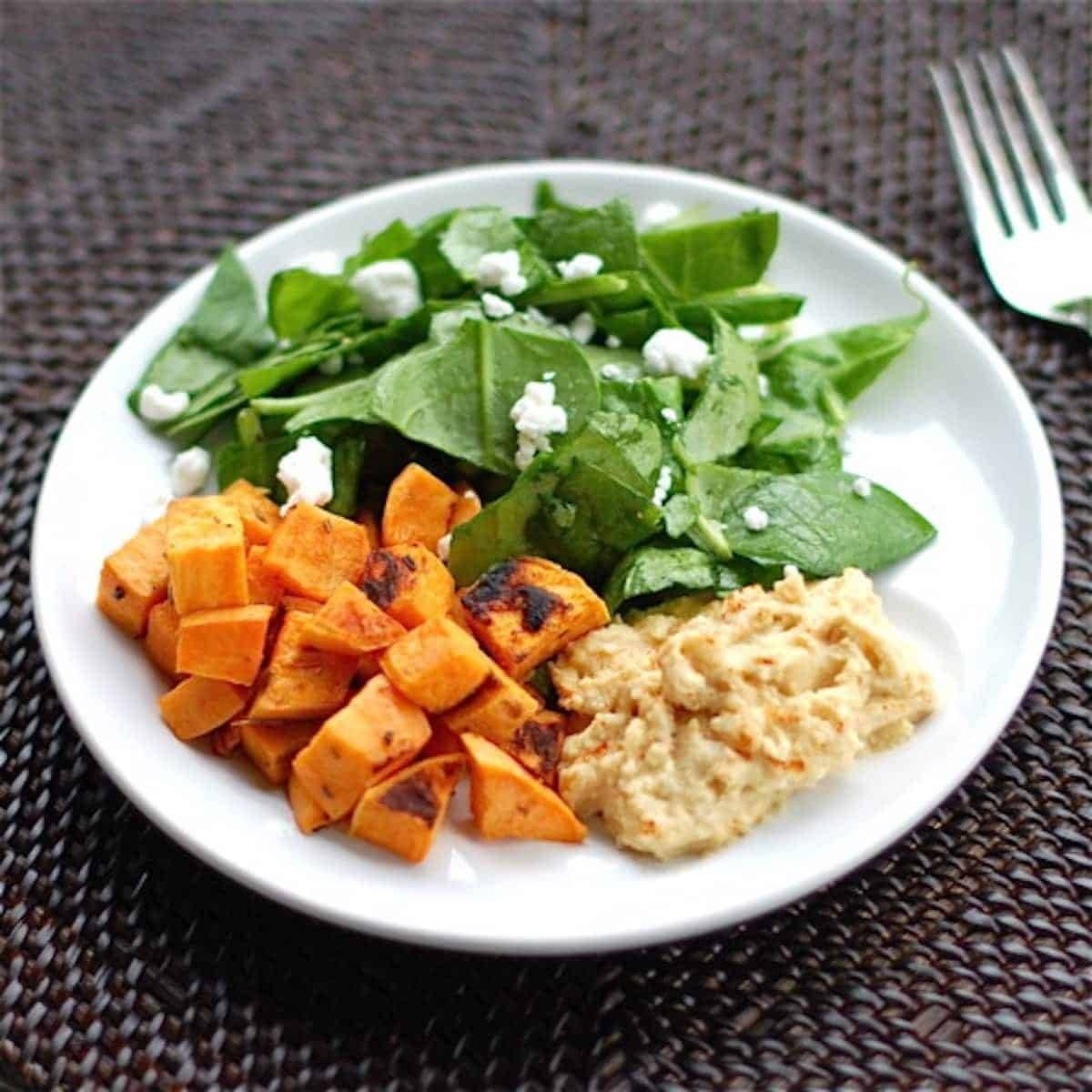 Roasted sweet potatoes with a side of hummus and a spinach salad on a white plate with a fork.